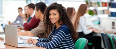 A Girl Sitting In A Library With Her Classmates And Starring At The Camera.