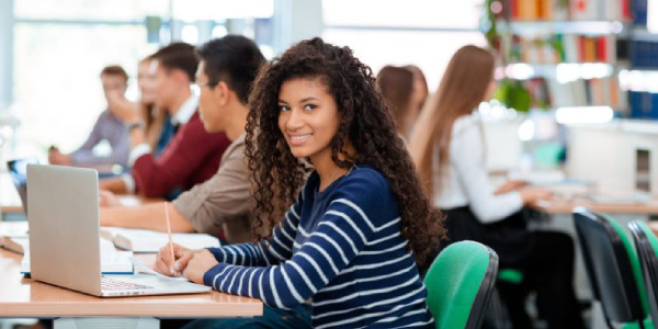 A Girl Sitting In A Library With Her Classmates And Starring At The Camera.