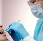A female dentist with specs oh ger eyes , head , nose , mouth and hand covered with masks and gloves performing a dental examination for a young femal patient.