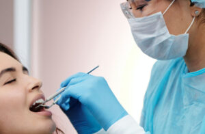 A female dentist with specs oh ger eyes , head , nose , mouth and hand covered with masks and gloves performing a dental examination for a young femal patient.