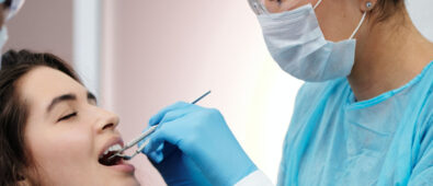 A female dentist with specs oh ger eyes , head , nose , mouth and hand covered with masks and gloves performing a dental examination for a young femal patient.