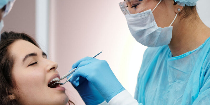 A female dentist with specs oh ger eyes , head , nose , mouth and hand covered with masks and gloves performing a dental examination for a young femal patient.