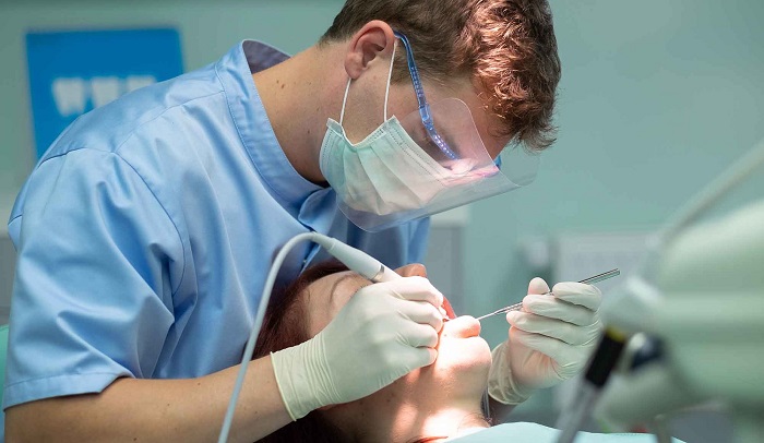 A male dentist with blue mask covering his mouth and nose examining the patient's teeth with dental equipments.