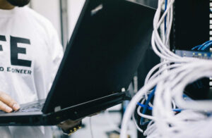 A field engineer is standing at the electrical wiring room with a laptop on his hand.