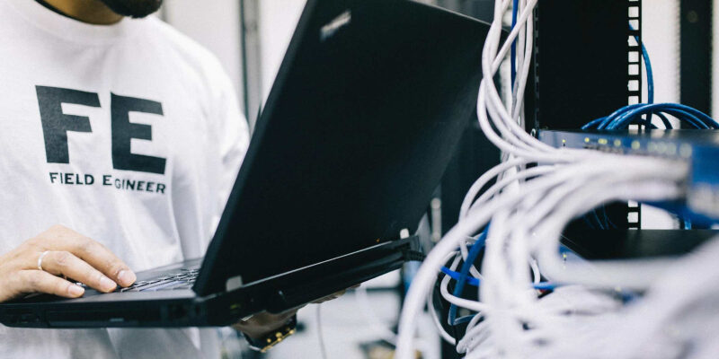 A field engineer is standing at the electrical wiring room with a laptop on his hand.